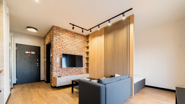 A modern living room with a brick fireplace, a grey sofa, wooden flooring, and a TV mounted on the wall. - Modena Jeżyce Deluxe Apartment (Poznań)