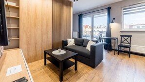 A modern living room with a sofa, a coffee table, a TV, and a view of the city through large windows. - Modena Jeżyce Deluxe Apartment (Poznań)