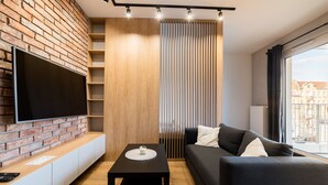 A modern living room with a brick wall, a flat-screen TV, a black sofa, a small table with a candle, and a view of buildings through a window. - Modena Jeżyce Deluxe Apartment (Poznań)