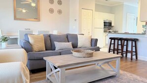 Un salon avec un canapé gris, une table basse en bois et une cuisine en arrière-plan.