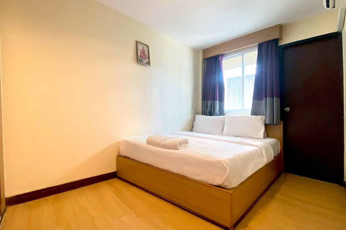 Un lit simple avec une couette blanche et une serviette enroulée, une fenêtre avec des rideaux, un petit tableau accroché au mur et un plancher en bois.