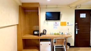 Una stanza compatta con una scrivania in legno, un piccolo frigorifero, una sedia, una televisione a parete e una porta con un cartello.
