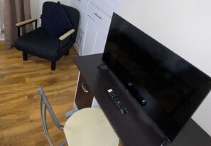 Une pièce avec un plancher en bois, un canapé noir, une chaise blanche, un téléviseur posé sur un meuble et une petite table avec une télécommande.