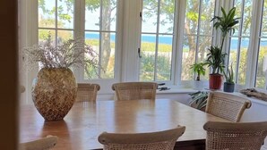 Un comedor con una mesa grande y sillas de mimbre, ventanas amplias con vista al mar, y plantas en macetas.