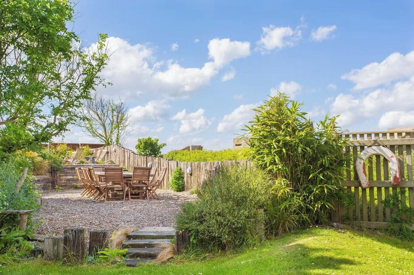 A rustic outdoor patio with a wooden table and chairs, surrounded by a wooden fence and lush greenery.