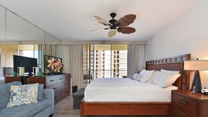 Een hotelkamer met een bed, een slaapbank, een televisie, een plafondventilator en een balkon met gordijnen.