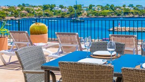 Un área de comedor frente al mar con sillas de mimbre, una mesa preparada para dos personas y vistas al océano y edificios a lo lejos.