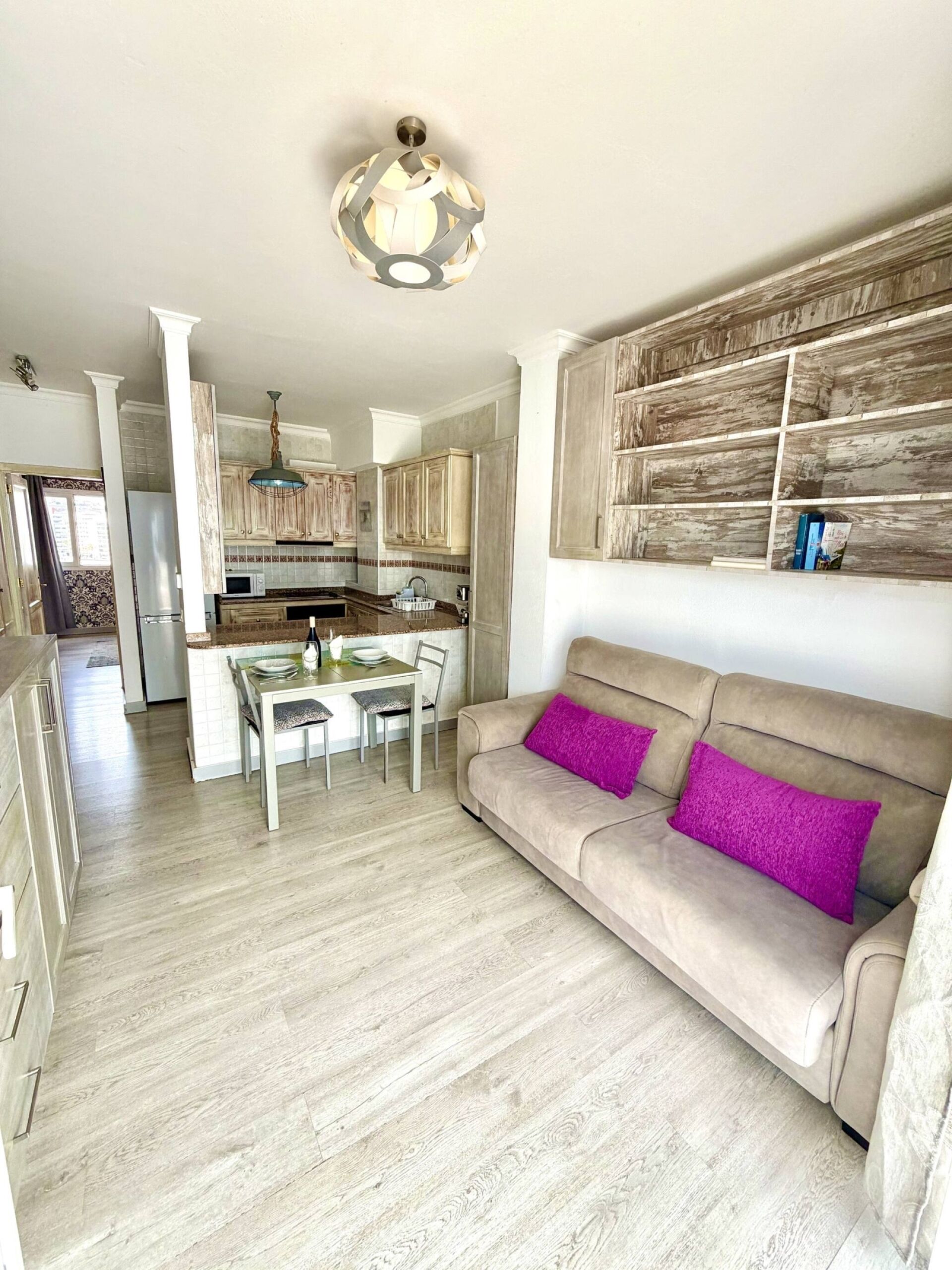 Una cocina moderna con armarios de madera, un comedor con mesa y sillas, y un sofá color beige con cojines morados.