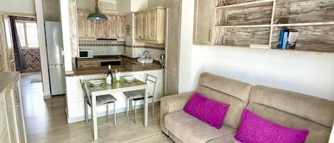 A modern kitchen with wooden cabinets, a dining area with a table and chairs, and a beige sofa with purple cushions.
