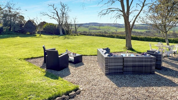 A grassy outdoor seating area with wicker furniture and a table, surrounded by trees and a view of the countryside. - Cedar Grange, Old Cleeve (Minehead)
