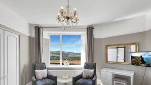 A bedroom with a large bed, two armchairs, a desk, a television, a chandelier, and a window with a view of the countryside. - Cedar Grange, Old Cleeve (Minehead)