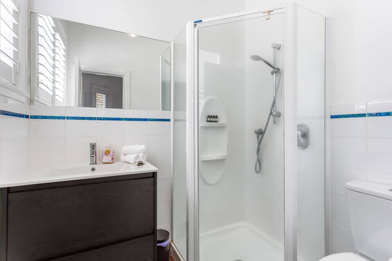 A modern bathroom with a glass shower enclosure, a white sink with a dark cabinet, and a mirror with a blue stripe.