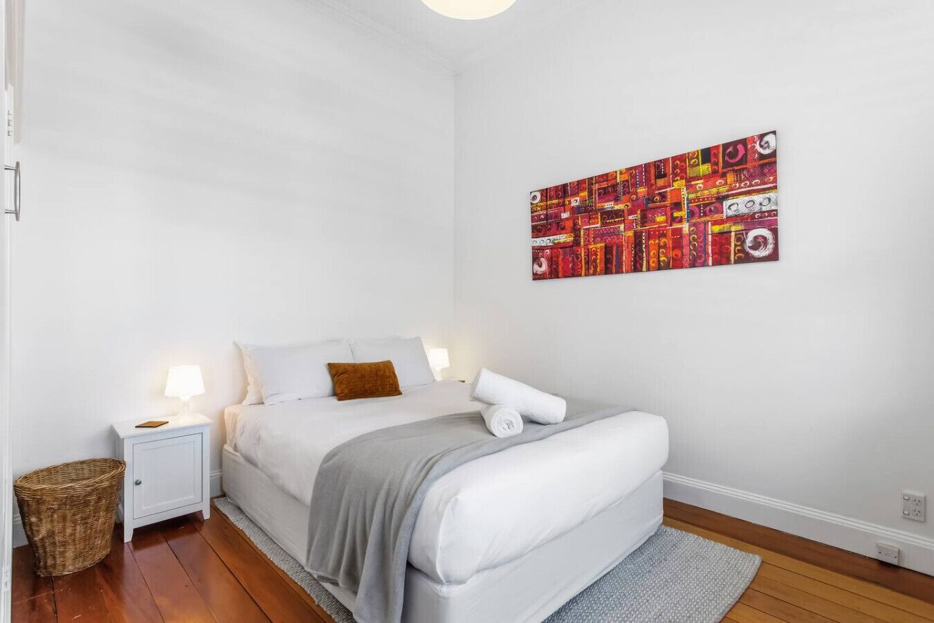 A bedroom with a bed, nightstand, lamp, and a colorful abstract painting on the wall.
