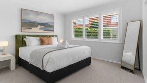 Un dormitorio con cama, mesita de noche, lámpara de noche, un cuadro, ventana con persianas y espejo.
