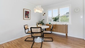 Un comedor con una mesa redonda, cuatro sillas, una lámpara colgante, una ventana con persianas y un reloj en la pared.