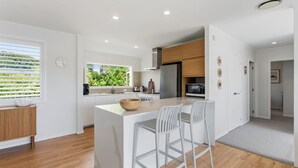 Una cocina moderna con una isla central, taburetes blancos y vistas a la vegetación por la ventana.
