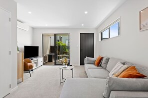 A modern living room with a grey sofa, a TV, and a glass door leading to an outdoor area.