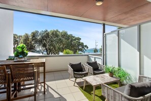 A balcony with a wooden table, wicker chairs, and a view of the sea. - Beach Front Bliss - by Furnished Rentals (Auckland)