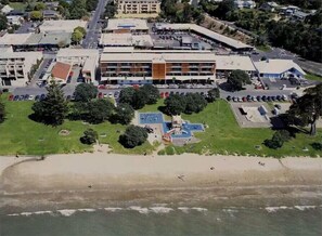 An aerial view of a coastal building complex with multiple structures, a parking area, and a beach in the foreground. - Beach Front Bliss - by Furnished Rentals (Auckland)