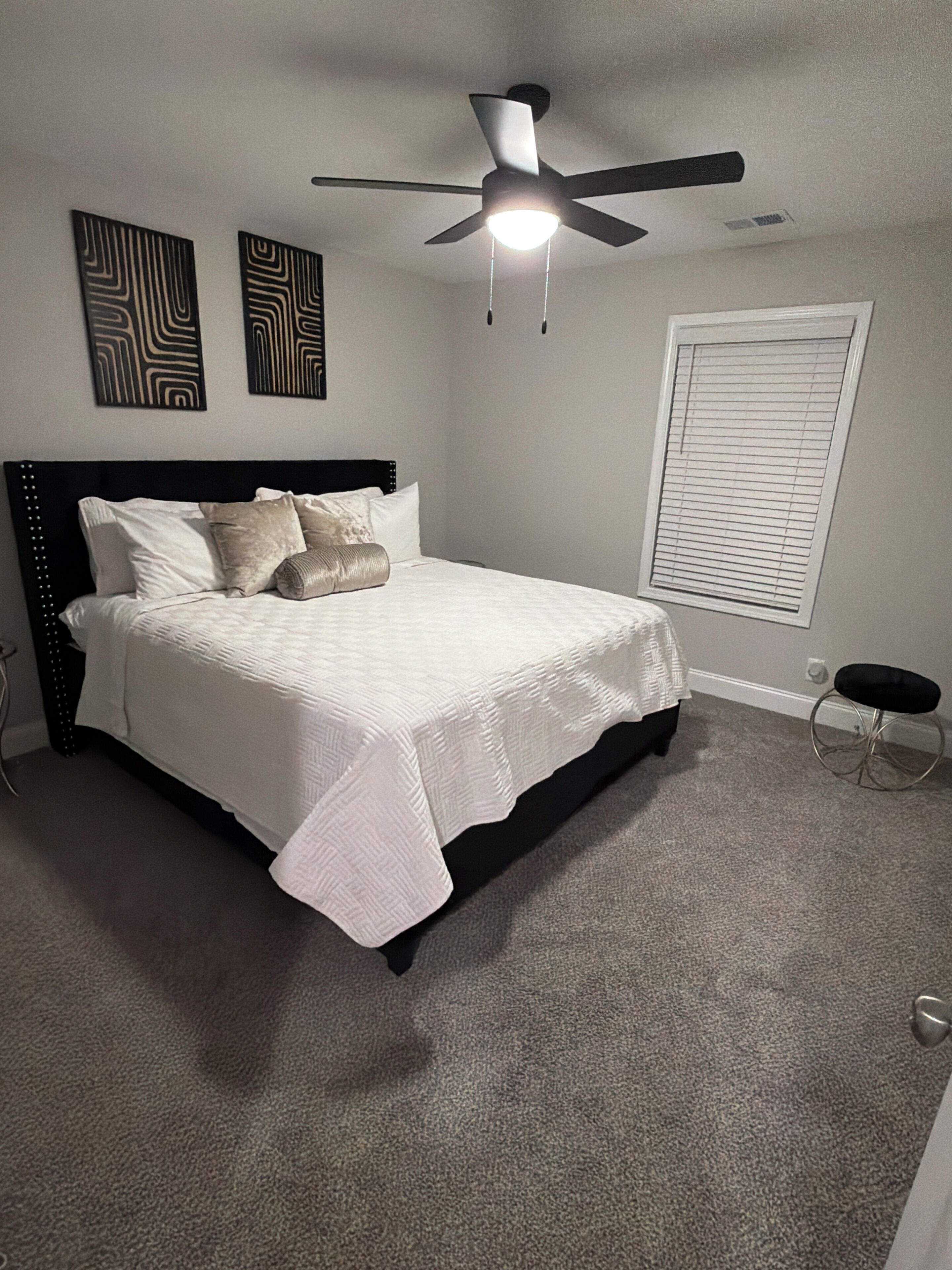 A bedroom with a bed, ceiling fan, artwork, window blinds, and a small stool.