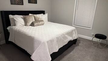 A bedroom with a bed, ceiling fan, artwork, window blinds, and a small stool.