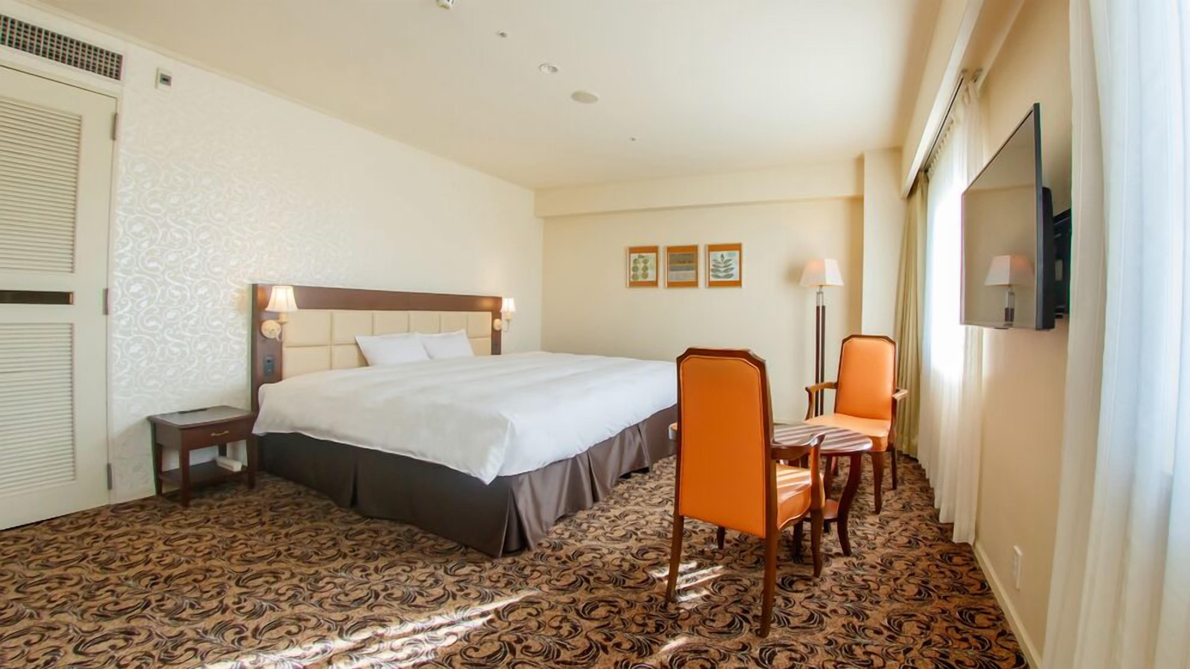 Quarto de hotel com uma cama grande, uma escrivaninha, uma cadeira laranja, televisão de tela plana e carpete estampado.