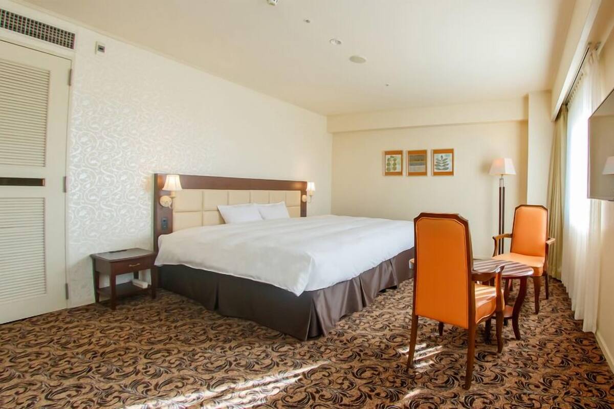 Une chambre d’hôtel avec un grand lit, un bureau, une chaise orange, un téléviseur à écran plat et une moquette à motifs.
