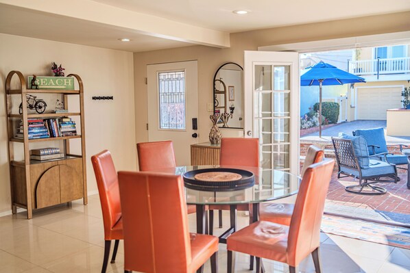 A dining area with a glass table surrounded by orange chairs, a bookshelf with books and a sign, and a view of the backyard with a blue umbrella. - Strand N Sea • Beach • Hot Tub • Pet Friendly • Channel Islands Harbor (Oxnard)