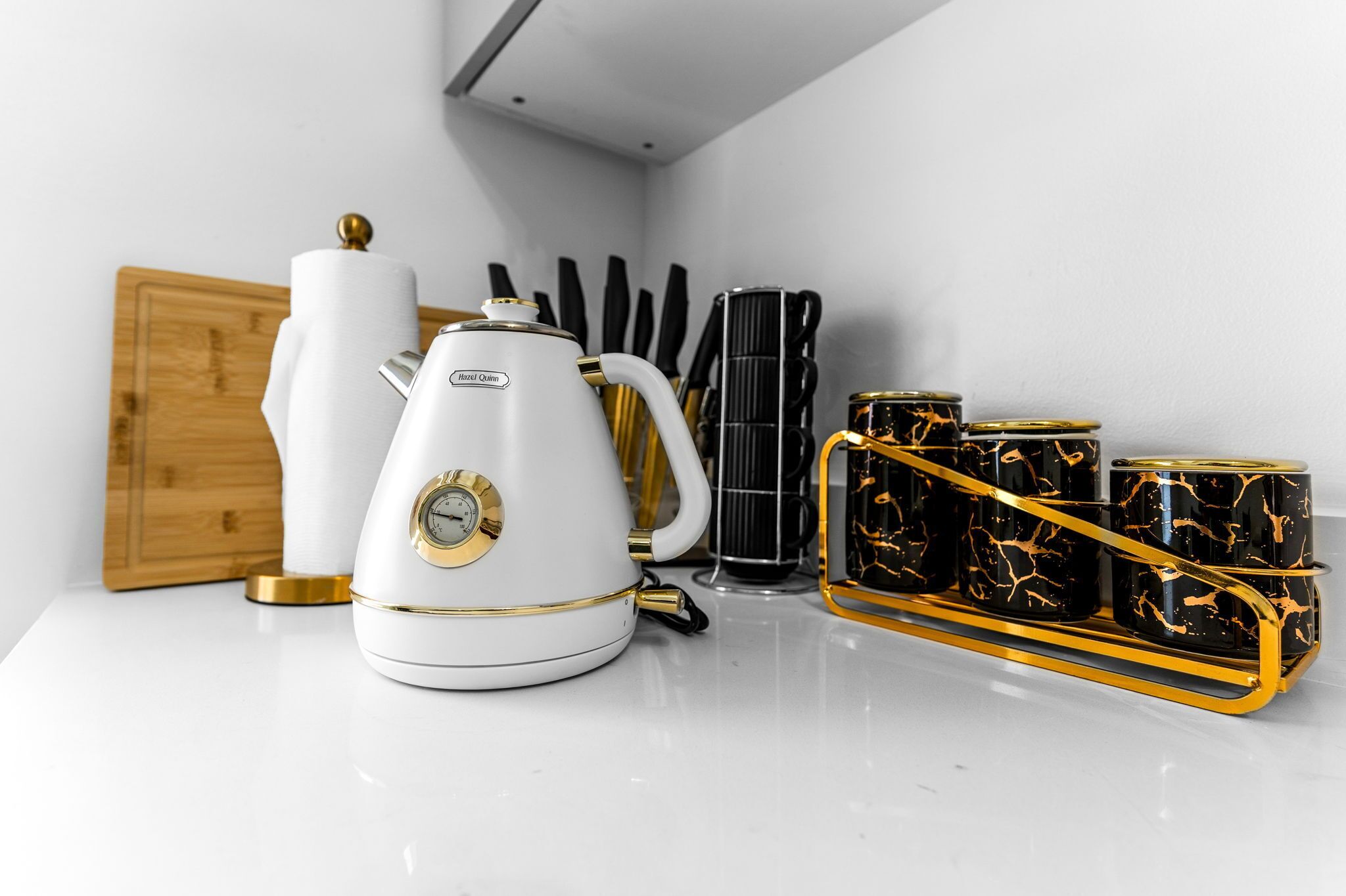 Une bouilloire blanche, une machine à café et un plateau avec des tasses noires et dorées, le tout posé sur un plan de travail blanc.