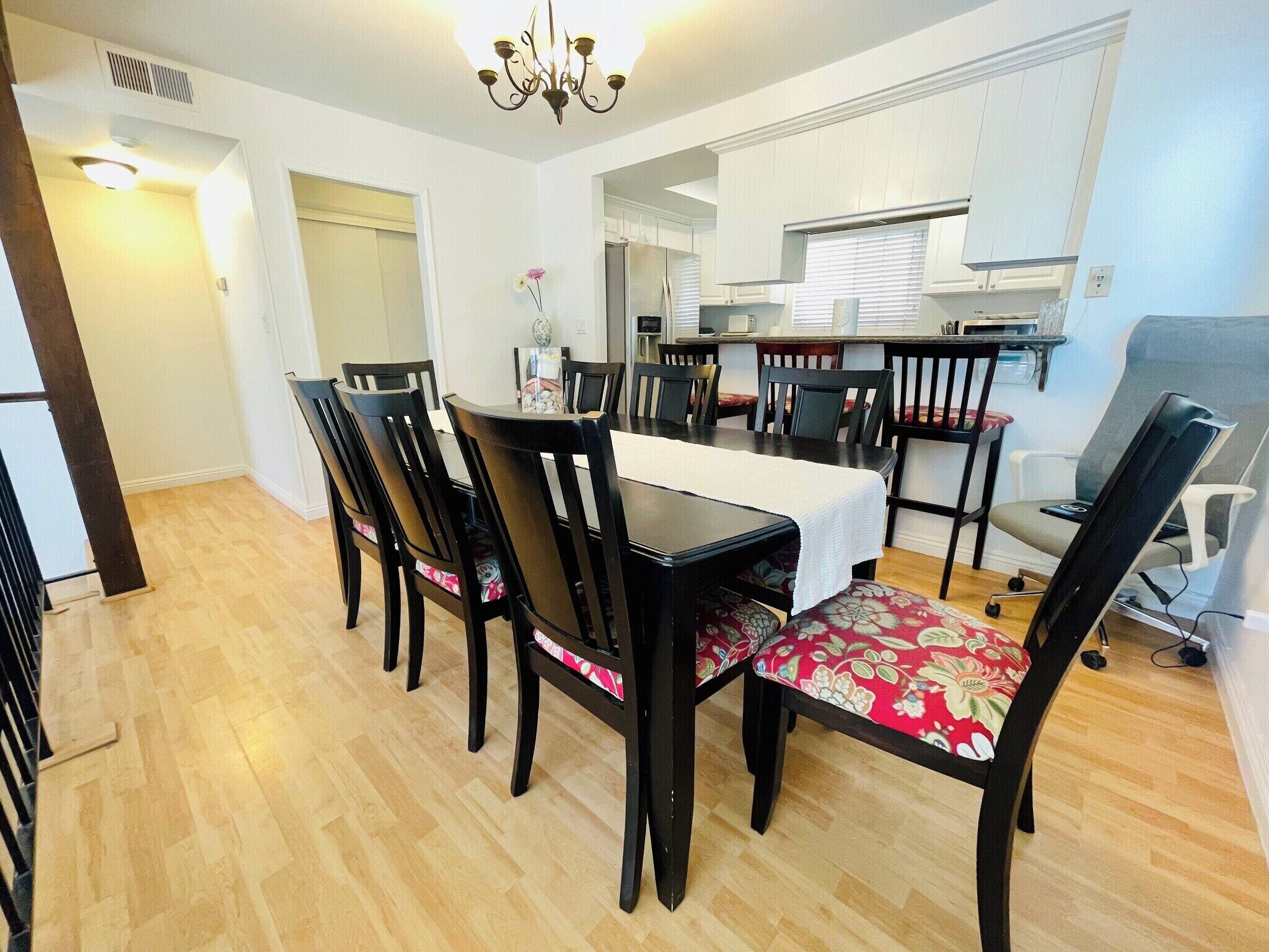A dining area with a table and chairs, a kitchen in the background, and a chandelier.