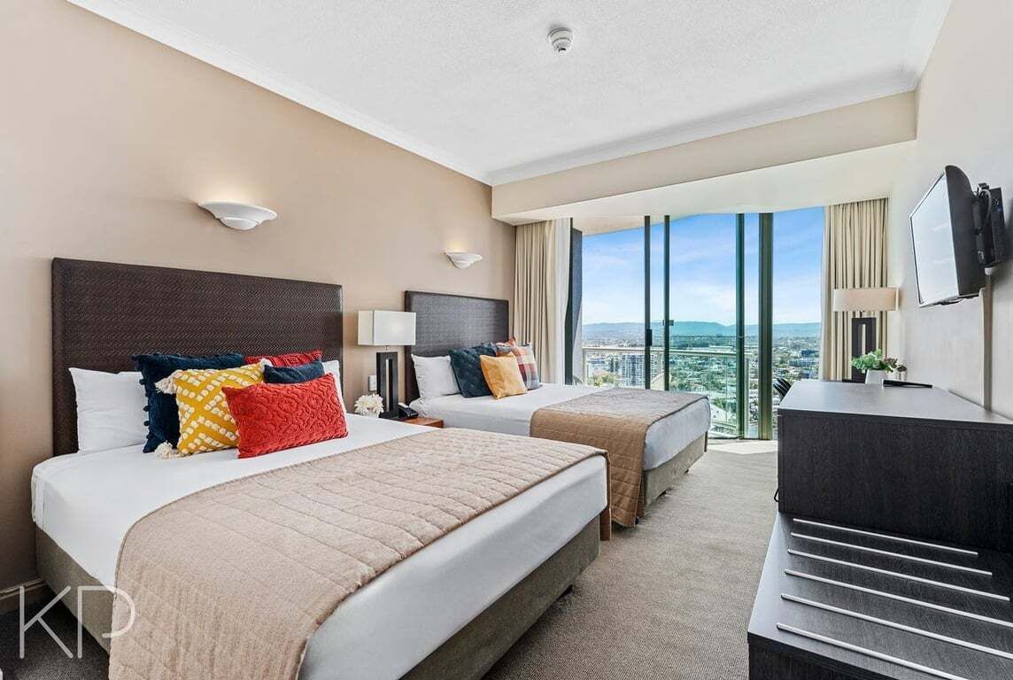 A hotel room with two beds, a desk, a TV, and a large window with a city view.