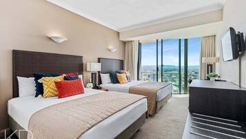 A hotel room with two beds, a desk, a TV, and a large window with a city view.