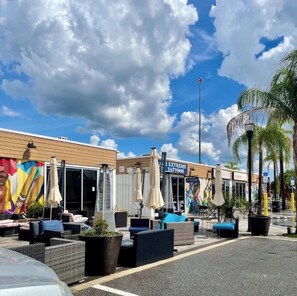 An outdoor seating area with umbrellas, a building with a tattoo shop, and palm trees. - Charming Quaint Getaway - Blocks from Downtown Zephyrhills - Pet Friendly (Zephyrhills)