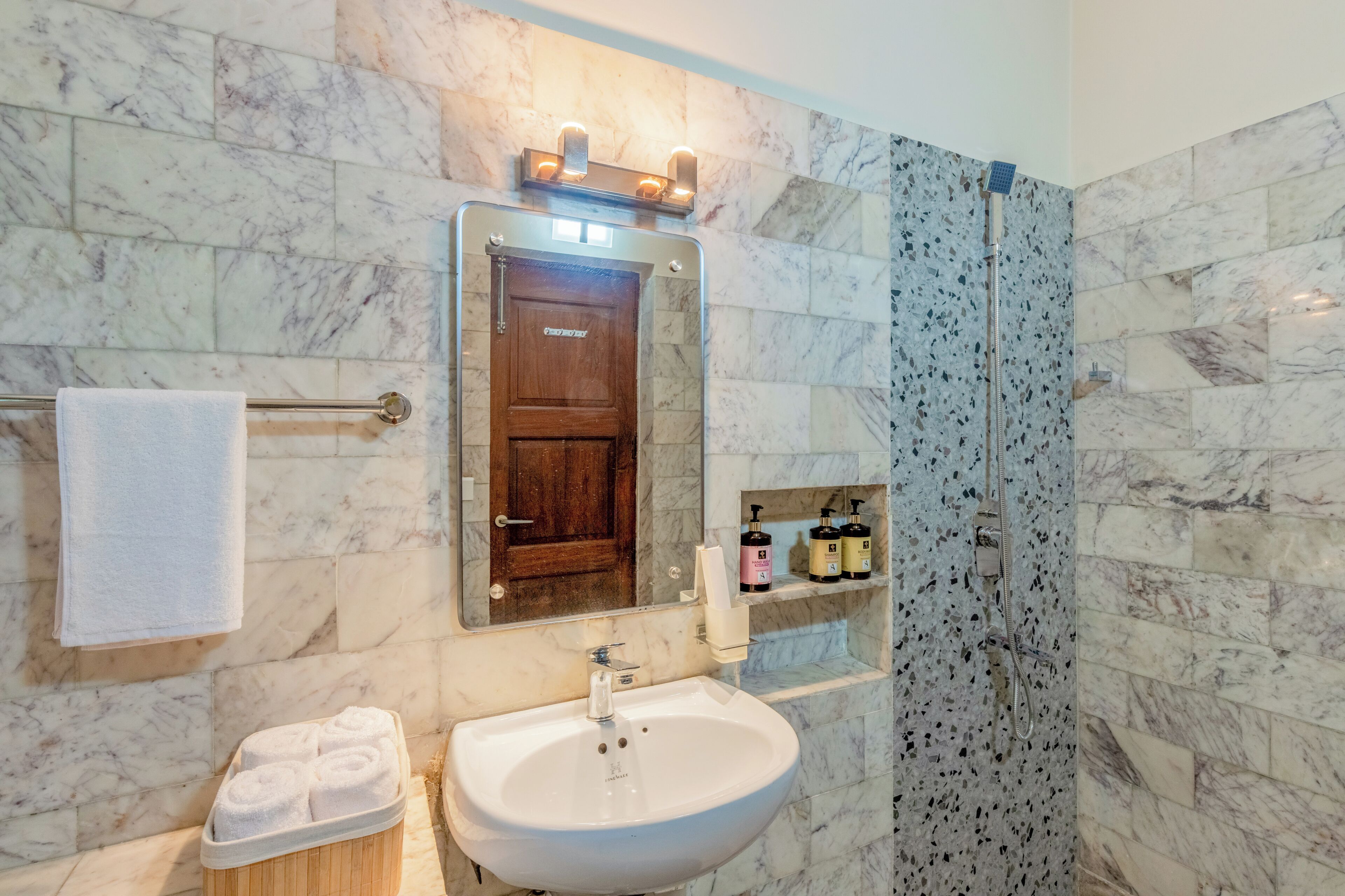 A bathroom with a marble wall, a sink, a mirror, a towel rack, a shower enclosure, and a shelf with toiletries.