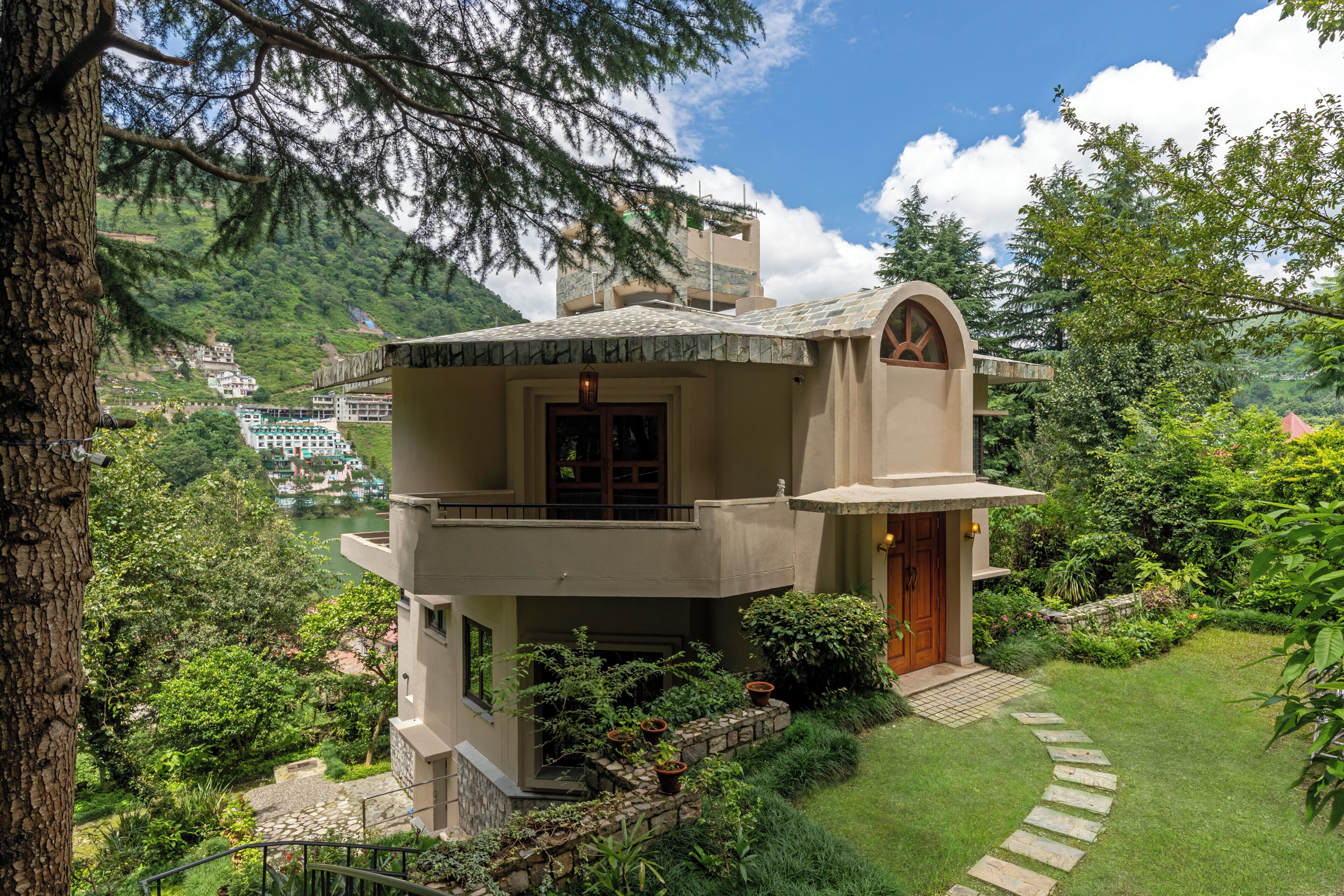 A two-story house with a stone pathway leading to the entrance, surrounded by lush greenery and a mountainous backdrop.