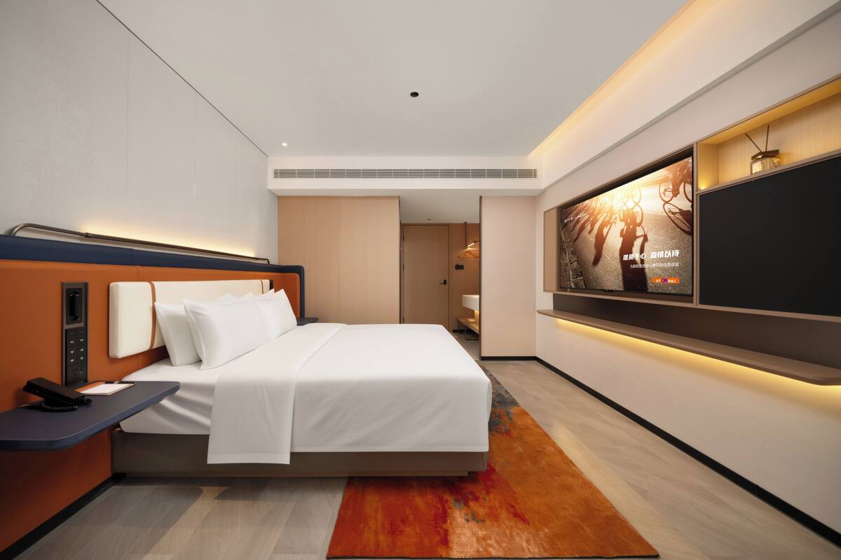 A modern hotel room with a large bed, a desk with a phone, a flat-screen TV mounted on the wall, and a small table with a lamp.