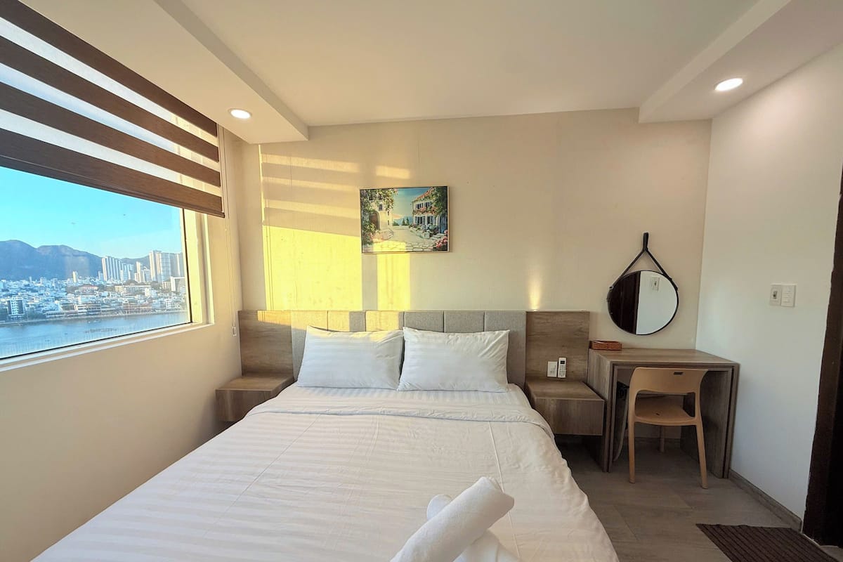 Quarto de hotel com cama, escrivaninha, cadeira, mesa de cabeceira, quadro e vista para a cidade.