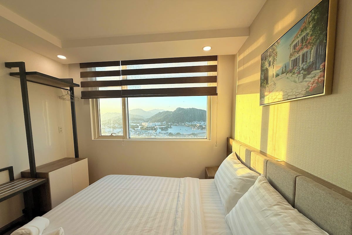 Quarto de hotel com cama, janela com vista para uma cidade e montanhas, um quadro na parede e cabeceira de madeira.