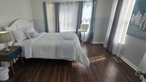 Una cama bien hecha con cabecera, una mesita de noche con lámpara, una silla y un cuadro en la pared.