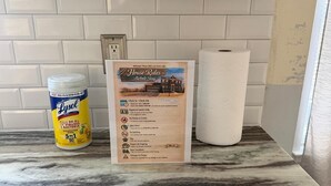 Una encimera de cocina con un spray desinfectante Lysol, un cartel de normas de la casa y un rollo de toallas de papel.