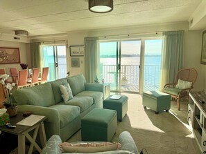 A living room with a sofa, ottoman, and a chair, a dining area with chairs and a table, and a balcony with a view of the sea. - Ocean City MD Harbour Island Bay View Condo (Ocean City)