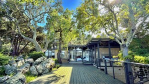 Una terraza de madera que conduce a un edificio moderno con paredes de vidrio, rodeado de árboles y rocas.