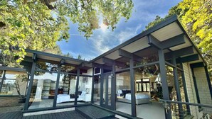 Una estructura moderna con paredes de vidrio y una terraza de madera, que incluye dos habitaciones con camas y mesitas de noche, y un baño a la vista.