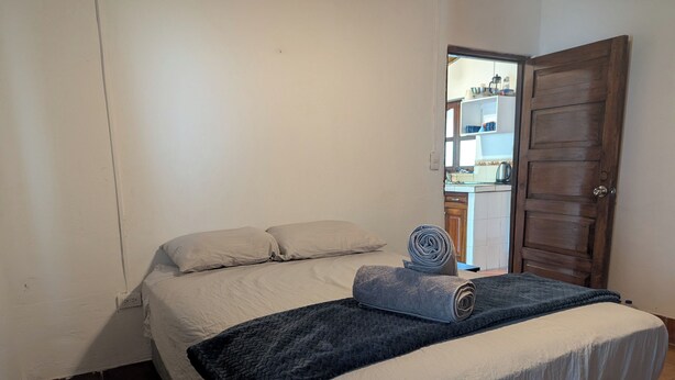 Un dormitorio con una cama, una toalla enrollada, un edredón oscuro, una puerta de madera y una cocina visible a través de una puerta abierta.