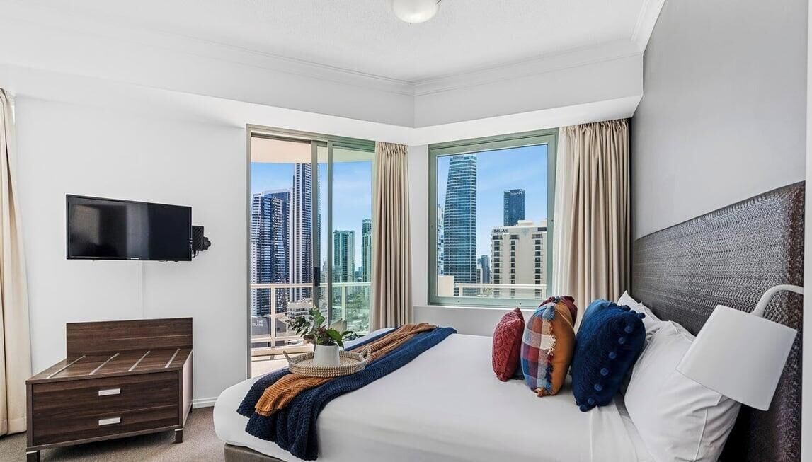 A hotel room with a bed, a TV, a dresser, and a balcony with a city view.