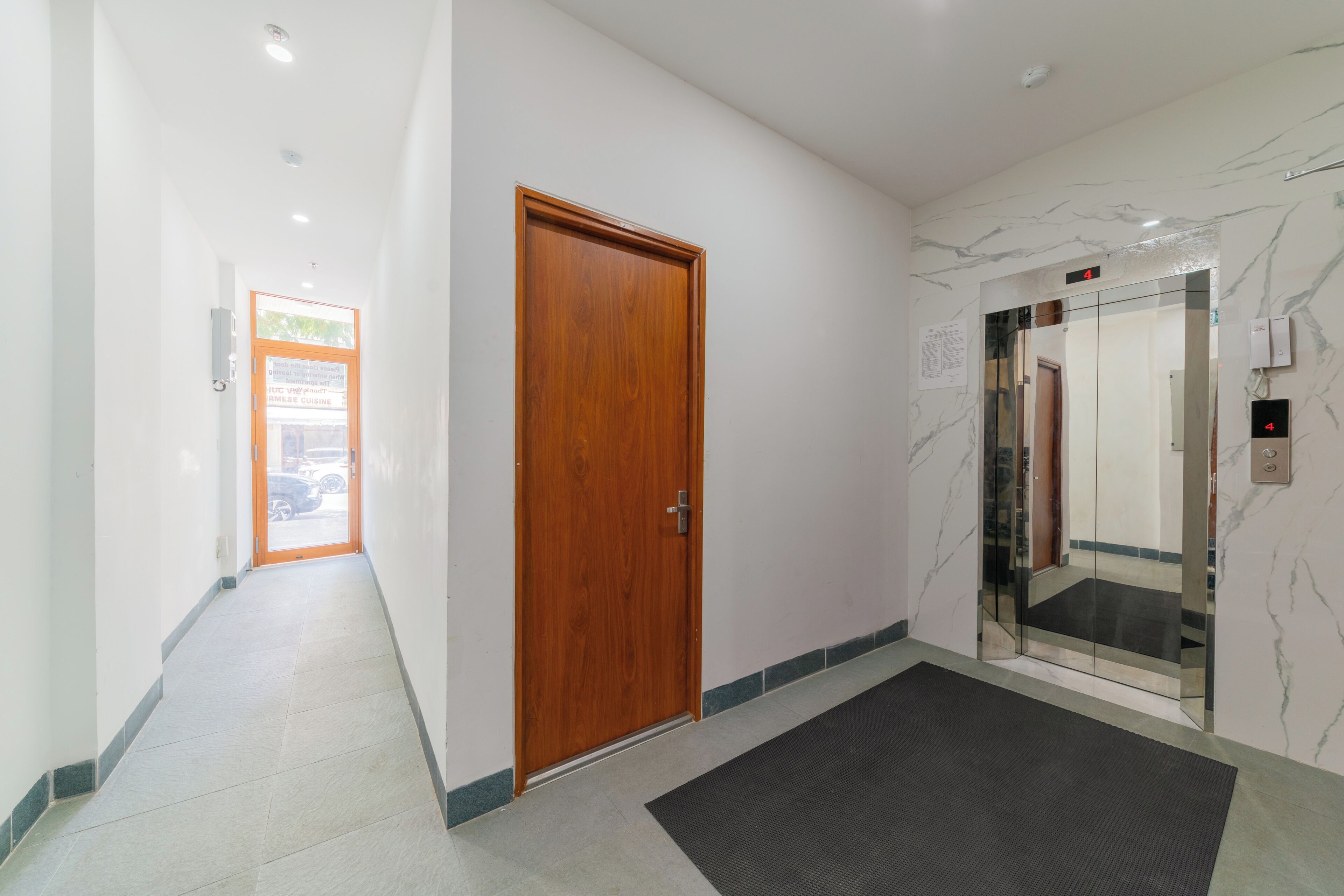 A hallway with a wooden door, a black mat, and an elevator with marble walls.
