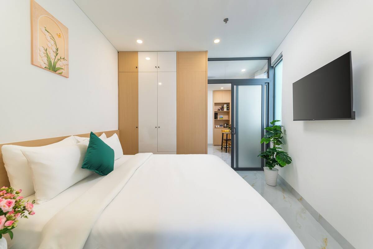 Ein modernes Hotelzimmer mit Bett, Flachbildfernseher, Blumentopf und Blick in den Küchenbereich.