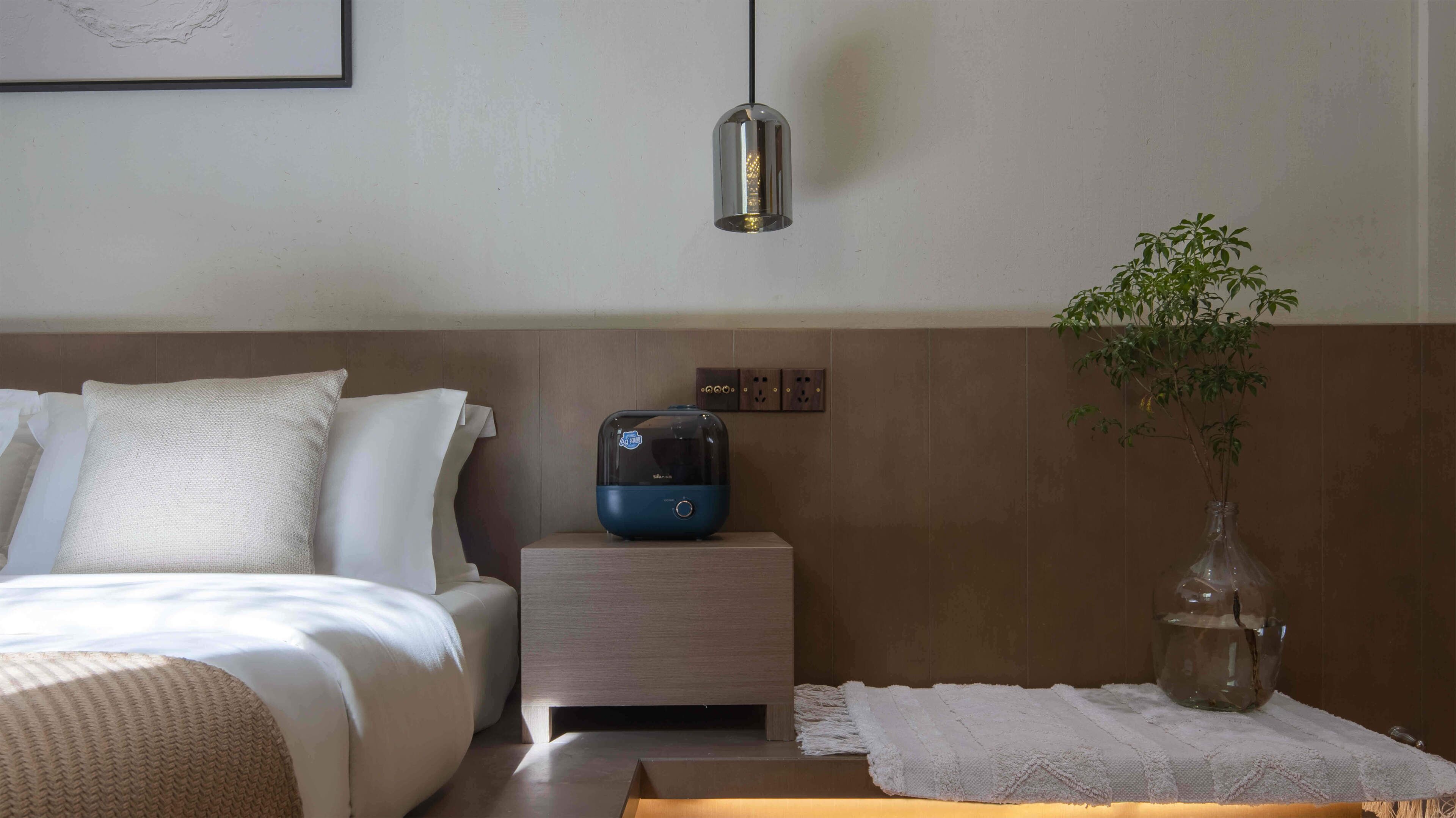 Een netjes opgemaakt bed met kussens, een nachtkastje met een blauw apparaat, een hanglamp en een kamerplant.
