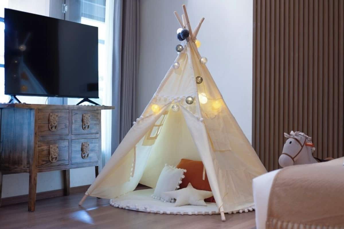 A cozy room with a bed, a TV, a wooden dresser, a hanging basket, and a teepee-style tent with lights.