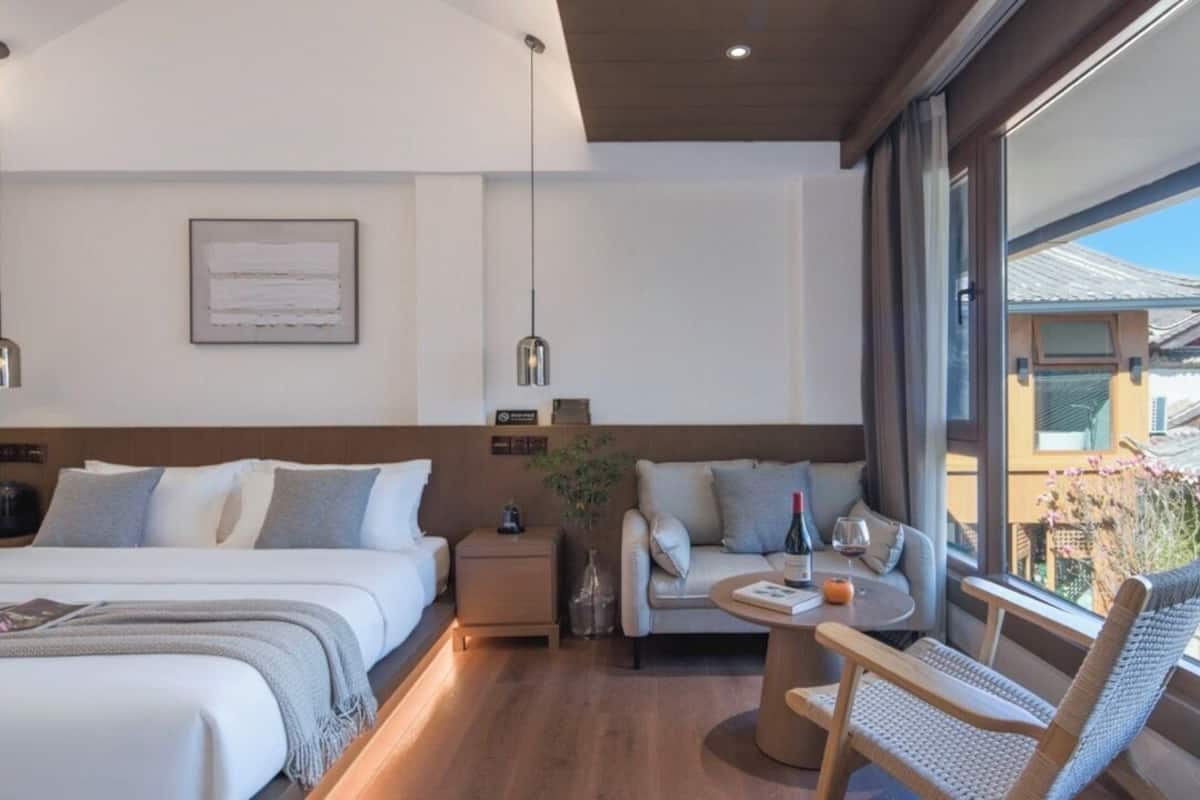 A modern hotel room with a large bed, a small sofa, a table with a bottle and glass, and a view of the outside.